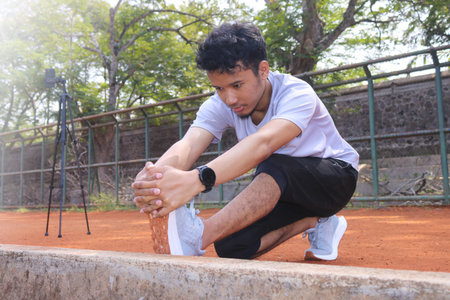 A man is stretching his leg on a dirt field. The man is wearing a white shirt and black shorts.