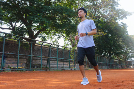 A man is running on a dirt field with a green fence behind him. The man is wearing a grey shirt and...
