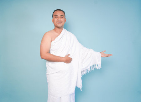 Asian friendly Muslim young man wearing ihram posing open arms, welcoming guests, over isolated blue...
