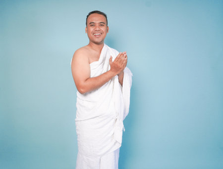 young Asian Muslim man wearing ihram posing praying and lifting head up over isolated blue...