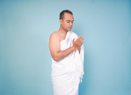 young Asian Muslim man wearing ihram posing praying and lifting head up over isolated blue...