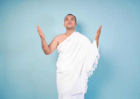 happy young Asian Muslim man wearing ihram gesturing, pose prayer pose raised, open hands, isolated...