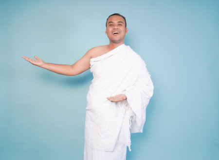 Asian friendly Muslim young man wearing ihram posing open arms, welcoming guests, over isolated blue...