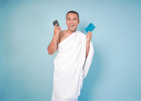 young Asian Muslim man wearing ihram doing video call pose while holding passport, isolated blue background. Hajj concept. islamの写真素材