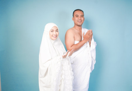 young Asian Muslim couple wearing ihram and prayer gown doing praying gesture over isolated by blue background. Hajj concept. concep islam muslim conceptの写真素材