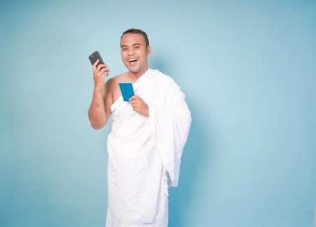 young Asian Muslim man wearing ihram doing video call pose while holding passport, isolated blue...