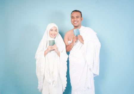 young Asian Muslim couple in ihram and prayer gown posing with a passport over isolated by blue background. Hajj concept.の写真素材