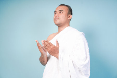 young Asian man Muslim wearing ihram and prayer gown doing praying gesture over isolated by blue background. Hajj concept. praying islam muslim conceptの写真素材