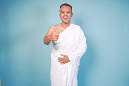 Excited Asian Muslim man wearing ihram gesture thumbs up pose, isolated blue background. Hajj...