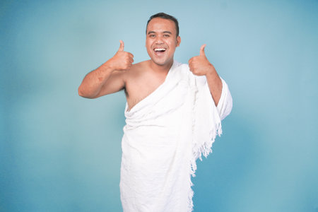 Excited Asian Muslim man wearing ihram gesture thumbs up pose, isolated blue background. Hajj...