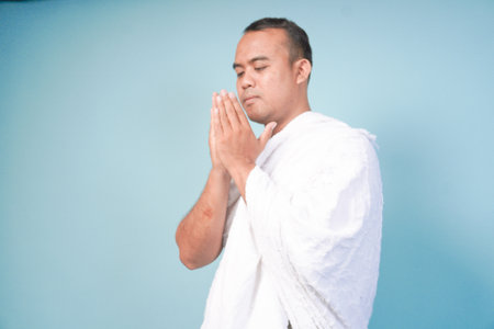 young Asian man Muslim wearing ihram and prayer gown doing praying gesture over isolated by blue background. Hajj concept. praying islam muslim conceptの写真素材