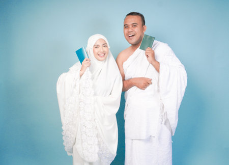 young Asian Muslim couple in ihram and prayer gown posing with a passport over isolated by blue background. Hajj concept.の写真素材