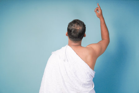 young Asian man Muslim wearing ihram and prayer gown doing praying gesture over isolated by blue background. Hajj concept. praying islam muslim conceptの写真素材
