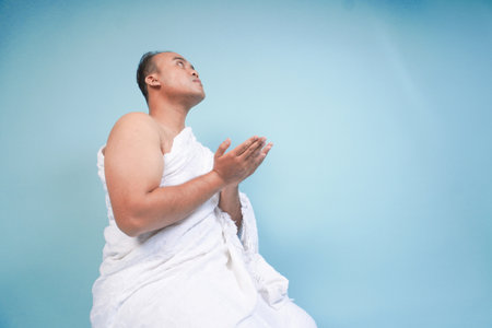Young serious Asian Muslim man wearing ihram sitting and praying holding hands up with hand gesture isolated on blue background. Hajj concept. islam muslim conceptの写真素材