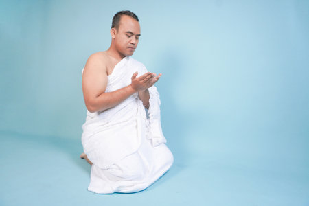 Young serious Asian Muslim man wearing ihram sitting and praying holding hands up with hand gesture isolated on blue background. Hajj concept. islam muslim conceptの写真素材