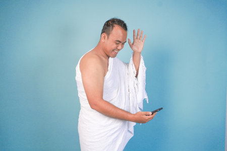 young Asian Muslim man wearing ihram doing video call pose while holding smarthphone, isolated blue background. Hajj concept. islamの写真素材