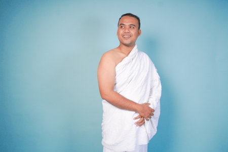 Asian Muslim young man wearing ihram posing how to use ihram on isolated blue background. Hajj...