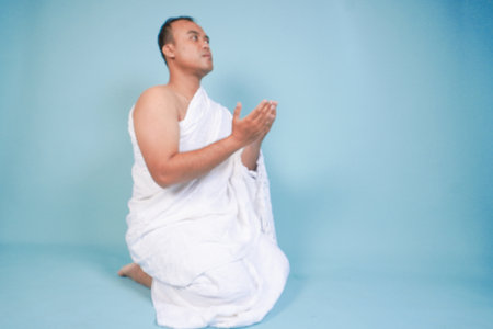 Young serious Asian Muslim man wearing ihram sitting and praying holding hands up with hand gesture...
