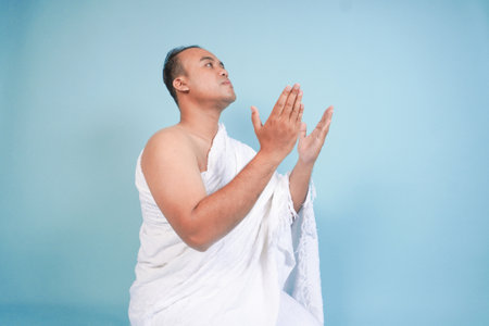 Young serious Asian Muslim man wearing ihram sitting and praying holding hands up with hand gesture isolated on blue background. Hajj concept. islam muslim conceptの写真素材