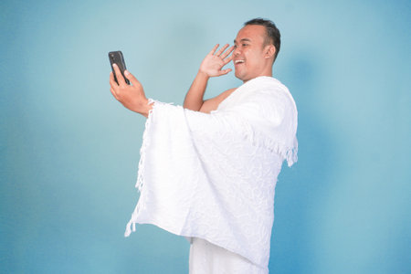 young Asian Muslim man wearing ihram doing video call pose while holding smarthphone, isolated blue background. Hajj concept. islamの写真素材