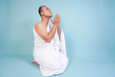 Young serious Asian Muslim man wearing ihram sitting and praying holding hands up with hand gesture isolated on blue background. Hajj concept. islam muslim conceptの写真素材