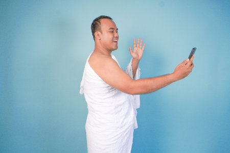 young Asian Muslim man wearing ihram doing video call pose while holding smarthphone, isolated blue background. Hajj concept. islamの写真素材