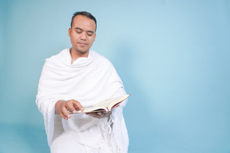 Serious young Asian Muslim man wearing ihram sitting praying worship reading al quran isolated on blue background. Hajj concept. Islamic Muslim concept. Hajj conceptの写真素材
