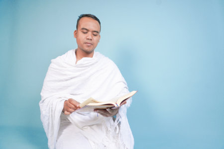Serious young Asian Muslim man wearing ihram sitting praying worship reading al quran isolated on blue background. Hajj concept. Islamic Muslim concept. Hajj conceptの写真素材