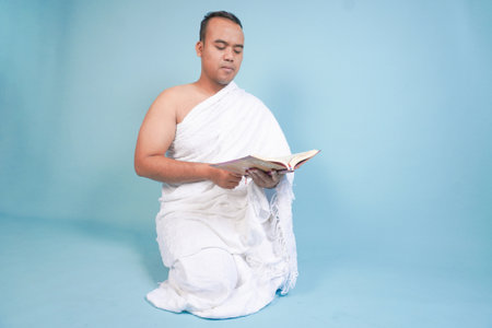 Serious young Asian Muslim man wearing ihram sitting praying worship reading al quran isolated on blue background. Hajj concept. Islamic Muslim concept. Hajj conceptの写真素材