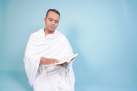 Serious young Asian Muslim man wearing ihram sitting praying worship reading al quran isolated on blue background. Hajj concept. Islamic Muslim concept. Hajj conceptの写真素材