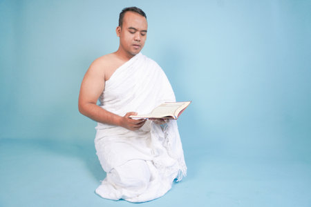 Serious young Asian Muslim man wearing ihram sitting praying worship reading al quran isolated on blue background. Hajj concept. Islamic Muslim concept. Hajj conceptの写真素材