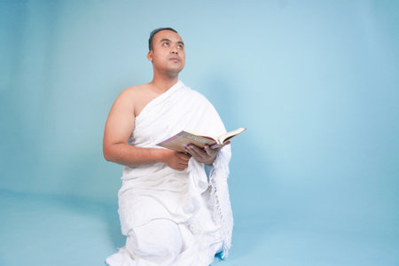Serious young Asian Muslim man wearing ihram sitting praying worship reading al quran isolated on blue background. Hajj concept. Islamic Muslim concept. Hajj conceptの写真素材