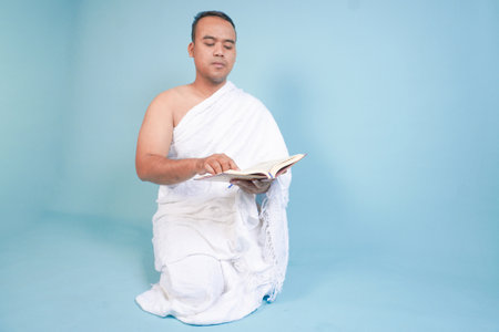 Serious young Asian Muslim man wearing ihram sitting praying worship reading al quran isolated on blue background. Hajj concept. Islamic Muslim concept. Hajj conceptの写真素材