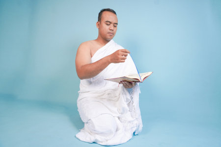 Serious young Asian Muslim man wearing ihram sitting praying worship reading al quran isolated on...