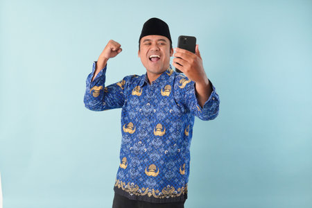 Smiling government worker man wearing kopri blue batik uniform He is doing video call while pointing to his smartphone. PNS, ASN civil servant concept.の写真素材