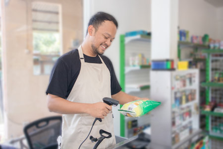 Smiling young Asian man as grocery store staff standing at cashier counter, cashier scanning grocery items and full of confidence in front of display shelvesの写真素材