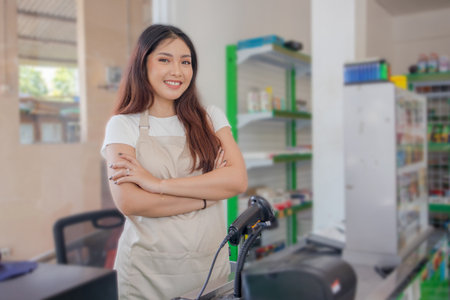 Smiling Asian young woman as groceries staff standing with crossed arms and full of confidence in front of display rackの写真素材