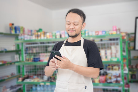 Asian male work as groceries store or modern market staff smiles and looks at the phone, wearing cream apron while standing in front of display rackの写真素材