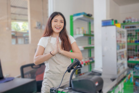 Young smiling Asian woman as grocery store staff standing at cashier counter with arms namaste pose and full of confidence in front of display shelvesの写真素材