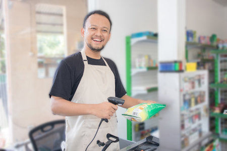 Smiling young Asian man as grocery store staff standing at cashier counter, cashier scanning grocery items and full of confidence in front of display shelvesの写真素材