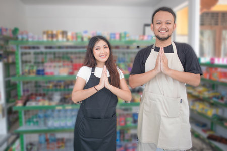 friendly Asian Cashier couple welcoming customers with open arms, namaste, cashier wearing black and beige apron standing in grocery store or convenience storeの写真素材