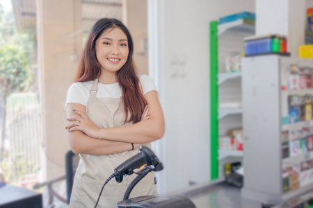 Smiling Asian young woman as groceries staff standing with crossed arms and full of confidence in front of display rackの写真素材