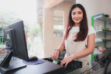 Smiling young Asian woman, grocery store staff wearing apron standing at cashier counter, returning...
