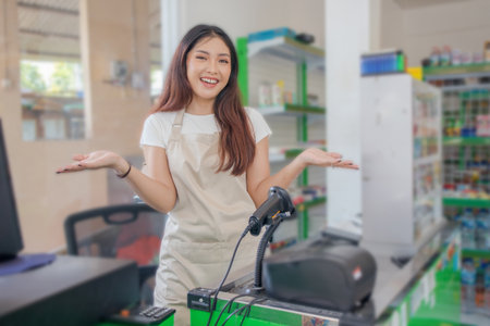 Young smiling Asian woman as grocery store staff standing at cashier counter with open hand gestureの写真素材