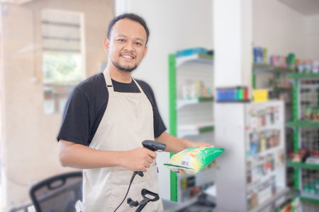 Smiling young Asian man as grocery store staff standing at cashier counter, cashier scanning grocery...