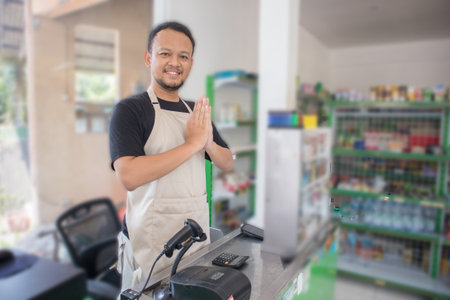 Young smiling Asian man as grocery store staff standing at cashier counter with arms namaste pose...
