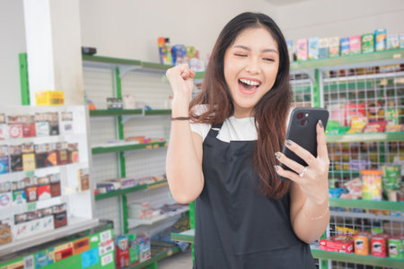 Asian female work as groceries store staff smiles and looks at the phone, wearing black apron while...