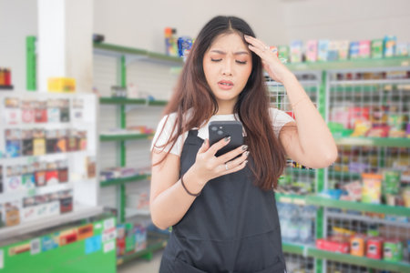 Asian female work as groceries store staff shocked, sad and looks at the phone, wearing black apron while standing in front of display rackの写真素材