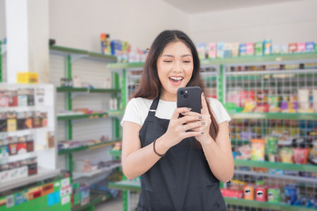 Asian female work as groceries store staff smiles and looks at the phone, wearing black apron while standing in front of display rackの写真素材