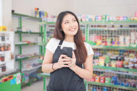 Asian female work as groceries store staff smiles and looks at the phone, wearing black apron while standing in front of display rackの写真素材